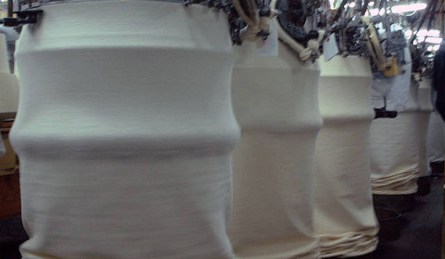 <strong>LOOPWHEELER｜ループウィラー</strong>　和歌山の工場のようす・カネキチ工業　「編まれた生地も重力によって下に下がっていき、床に用意されたタライのような丸いトレイの中に重なっていく仕組みです。かたや現代の高速編み機だと、遅いものでもその10倍、早いもので30倍。もちろん目で追える早さではありません」