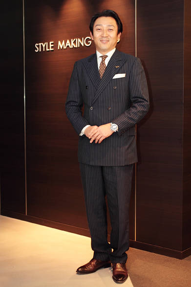 ネイビーストライプのダブルブレストに、明るめのブラウンのネクタイで登場