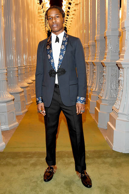 <strong>A$AP Rocky（エイサップ・ロッキー）</strong><br /> ウェア：2017年クルーズコレクションよりルック39のコントラスト プリントのミッドナイトブルー ウール ショールラペル ワンボタン タキシード、ホワイト ドレス シャツ、タイガーヘッド ブローチ付きグログラン リボン タイ<br /> シューズ：ファー付 スリッパ